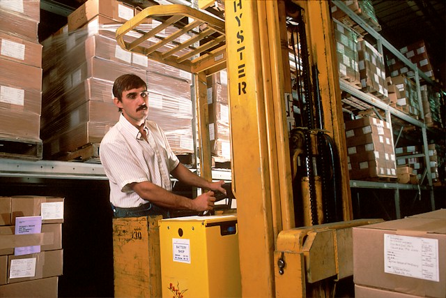 A man with a moustache, operating a standing forklift , with cardboard boxes behind him stacked high