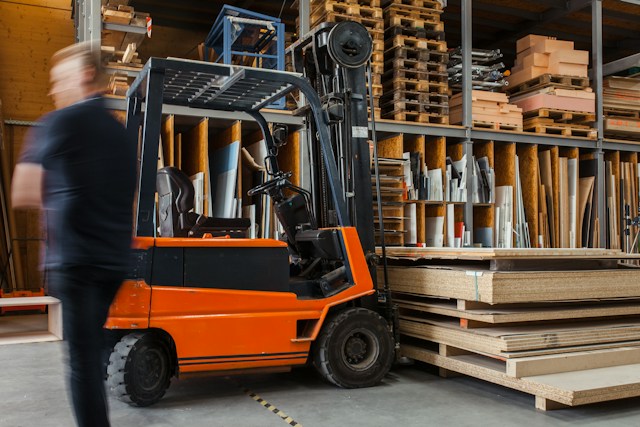 A forklift, with its prongs under a pallet of wood, inside a warehouse