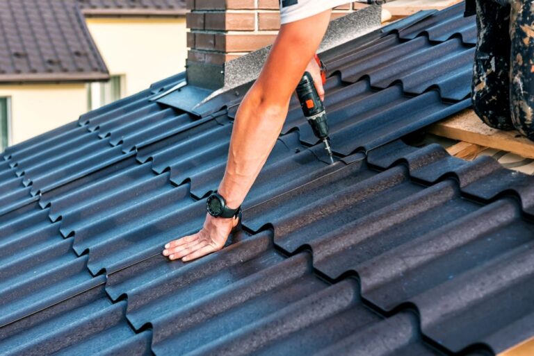 A roofer installing dark metal roofing sheets with a power drill on a residential roof.