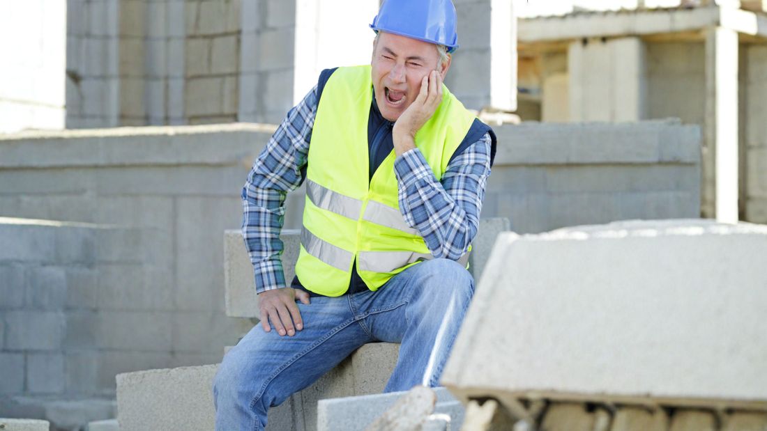 A construction worker in a high-vis vest and hard hat sitting on concrete blocks, holding his jaw in pain from a toothache on the worksite.
