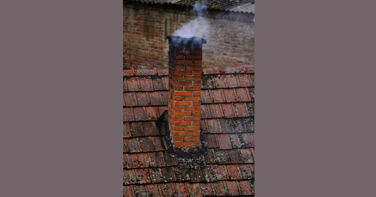 Smoke rising from an old brick chimney on a tiled roof, highlighting the need for regular chimney sweep services in Wellington.
