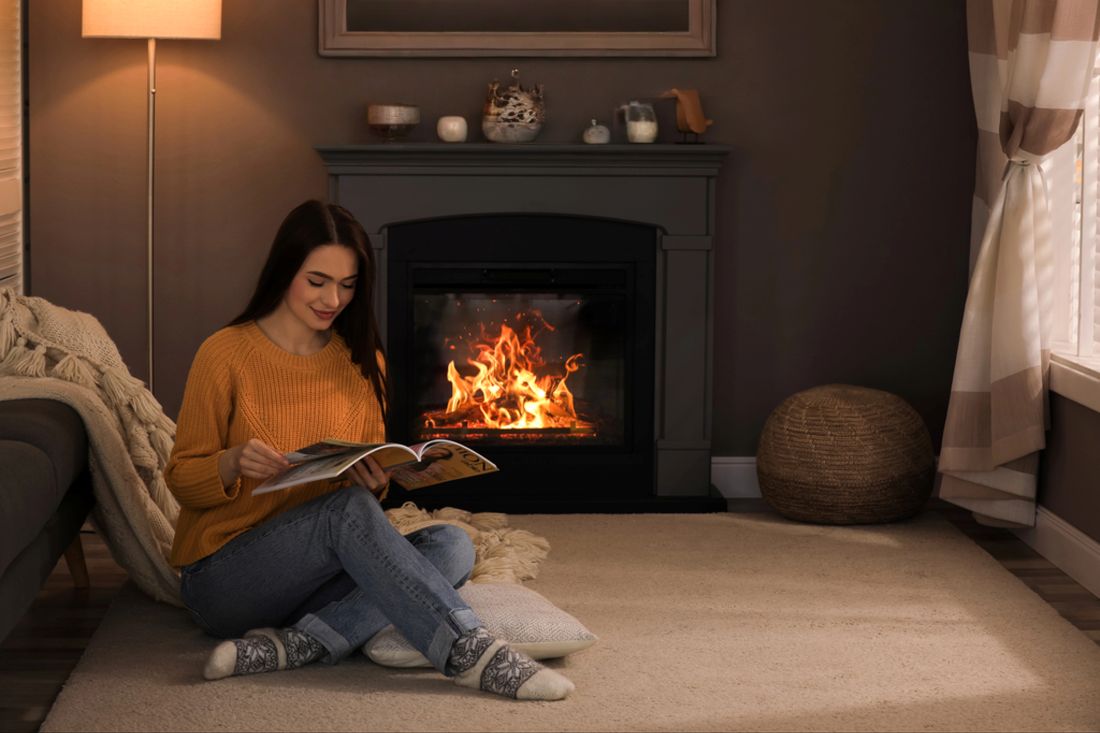 A woman reading a magazine while sitting on a cosy carpet in front of a lit fireplace, signifying the warmth and comfort of home heating in Wellington.
