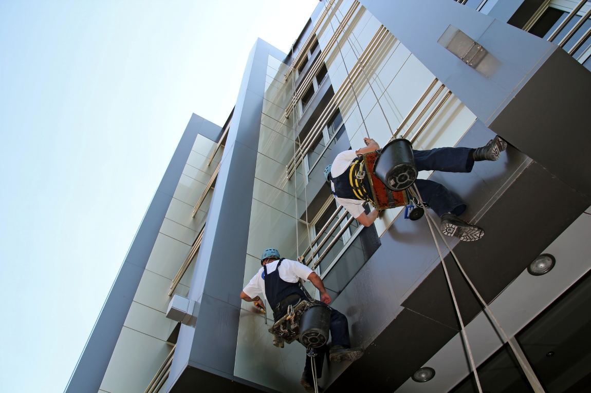 Professional cleaners using safety harnesses while washing a multi-storey commercial building exterior, showing proper technique and safety standards for expert building washing services.