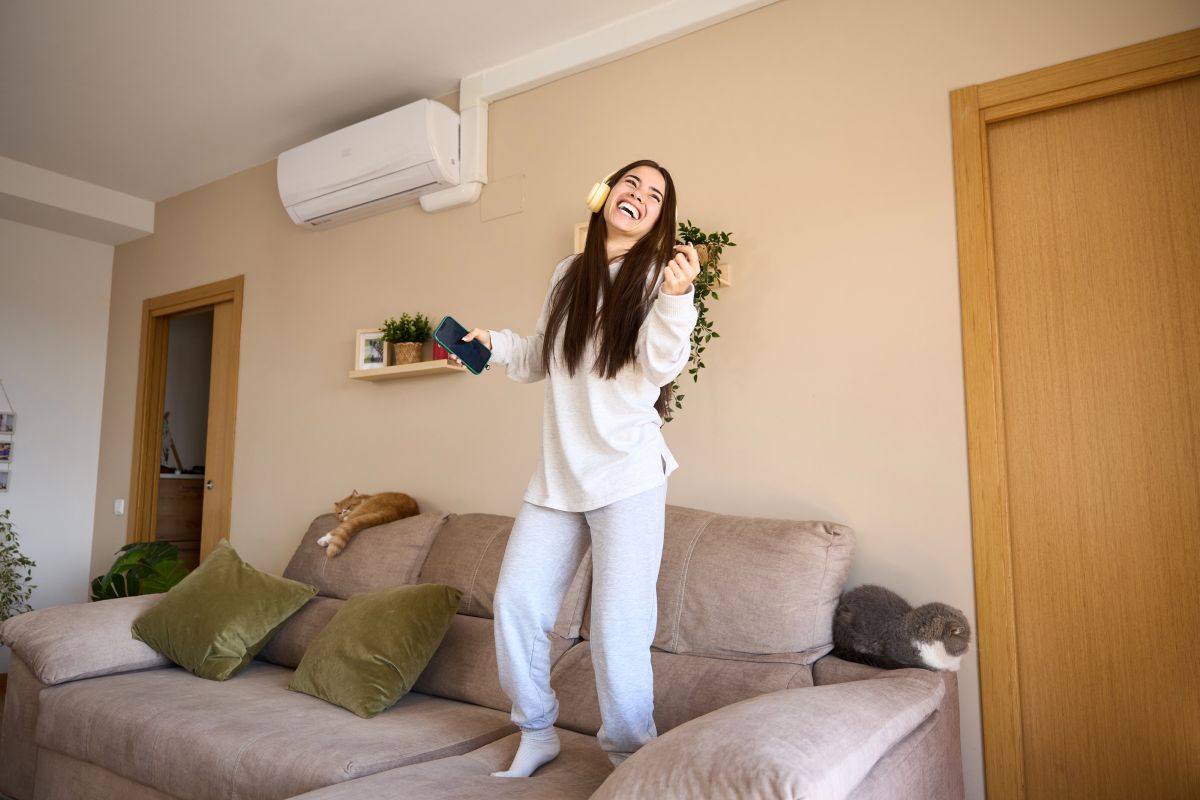 A young woman dancing on a couch in her living room, smiling and listening to music with headphones, while a heat pump is mounted on the wall providing warmth. Two cats are resting on the couch.